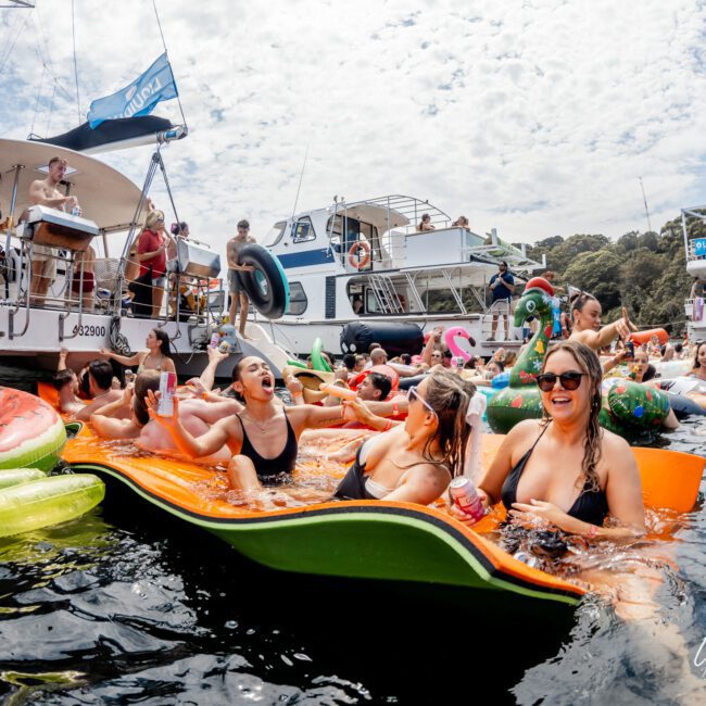 A lively group of people in swimsuits relax and laugh on large pool floats in the water, surrounded by boats and more partygoers under a partly cloudy sky.