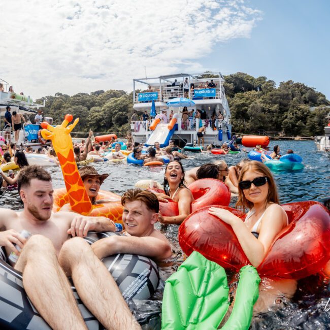 A group of people float in colorful inflatable pool toys on the water near boats, smiling and enjoying a lively party under a partly cloudy sky. Other groups are seen celebrating on the boats and in the water nearby.