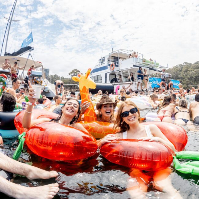 People relax on colorful pool floats in the water near yachts during a lively boat party, smiling and enjoying the sunny day with drinks and inflatable toys. Trees and a partly cloudy sky are visible in the background.