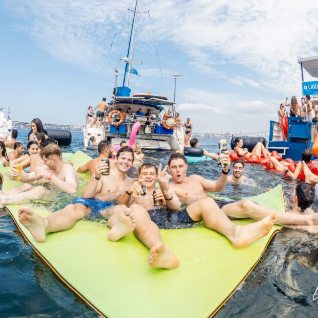 A group of young adults relax on a floating mat in the ocean, holding drinks and smiling. Behind them, more people enjoy the water and boats under a partly cloudy sky, creating a lively party atmosphere.