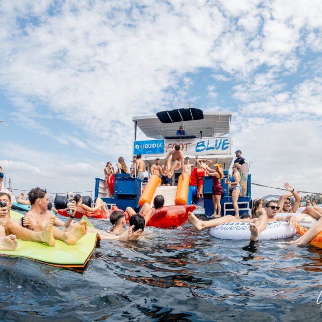 A group of people enjoy a pool party on the water, relaxing on colorful inflatables and slides next to a boat under a partly cloudy sky. The atmosphere is lively and fun.