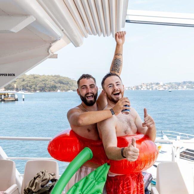 Two smiling men in swim trunks and inflatable rings pose playfully on a boat deck, with one giving a thumbs-up. There is water and a tree-lined shore in the background on a sunny day.