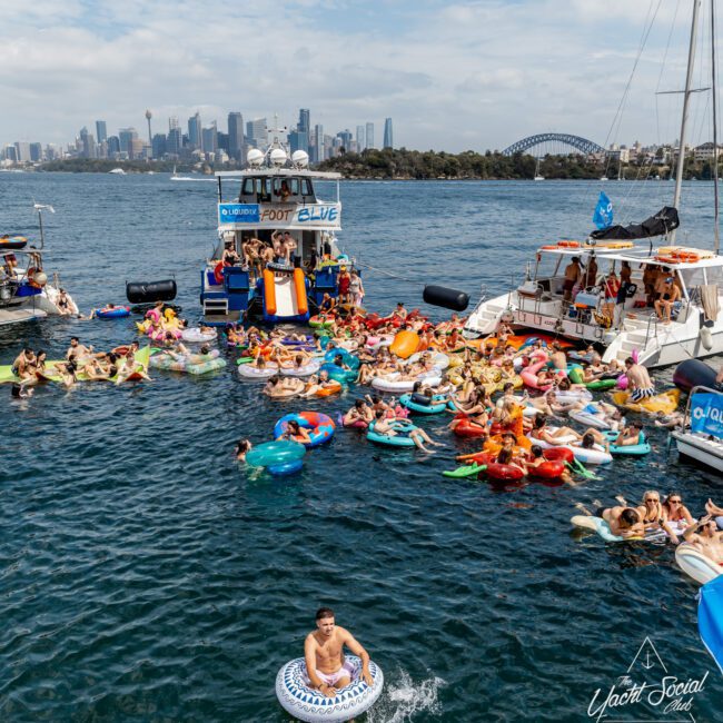 A large group of people in colorful inflatables enjoy a lively pool party in the water near boats, with a city skyline and Sydney Harbour Bridge visible in the background under a clear sky.