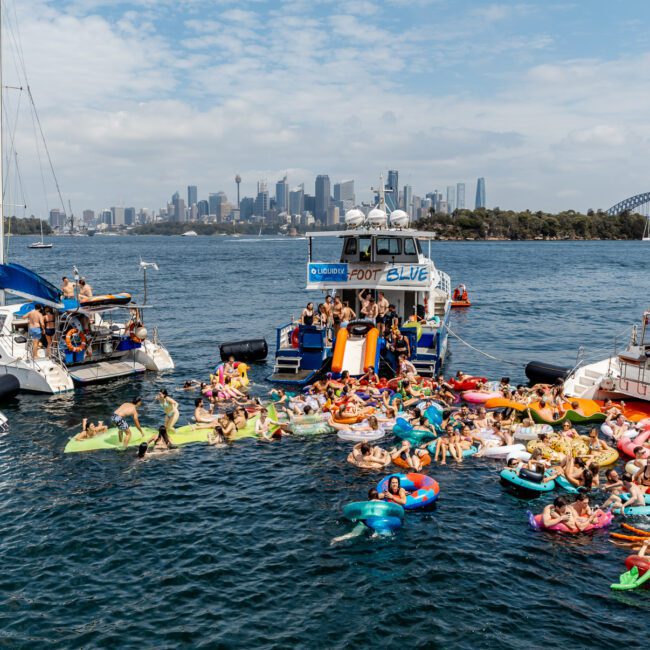 A lively group of people on colorful inflatables relax and socialize in the water between boats, with Sydney's city skyline and the Harbour Bridge visible in the background on a sunny day.
