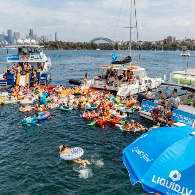 A lively group of people on colorful pool floats gather near boats on the water, with Sydney’s Harbour Bridge and city skyline in the background. A swimmer in an inflatable ring heads towards the crowd. A blue Liquid I.V. umbrella is visible.