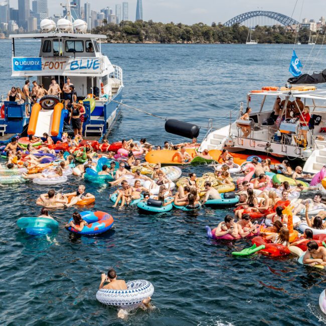 A large group of people on colorful inflatable rafts and floaties gather in the water between two boats, with a city skyline and the Sydney Harbour Bridge visible in the background.