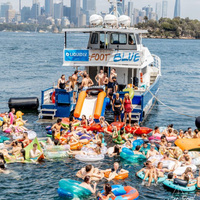 A lively crowd on colorful inflatables floats in the water near a boat labeled "Foot Blue," with people on the deck and a city skyline in the background on a sunny day.