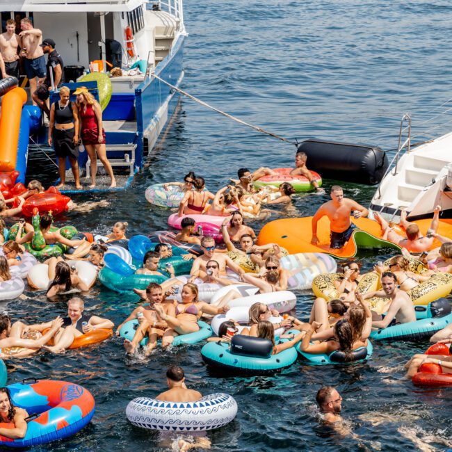 A large group of people relax on colorful inflatable floats in the water near a docked boat on a sunny day, enjoying a lively party atmosphere.