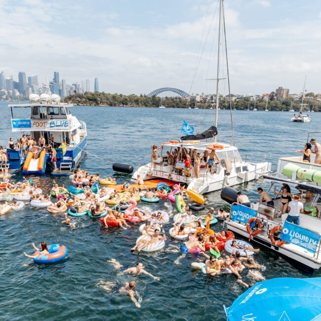 A lively crowd of people on colorful inflatables float in the water near boats, with Sydney's skyline and the Harbour Bridge visible in the background under a partly cloudy sky.