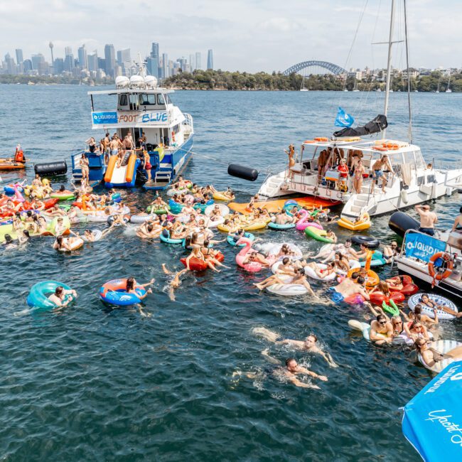 A large group of people on colorful inflatables and pool floats gather in the water between boats during a lively party; city skyline and Sydney Harbour Bridge are visible in the background.