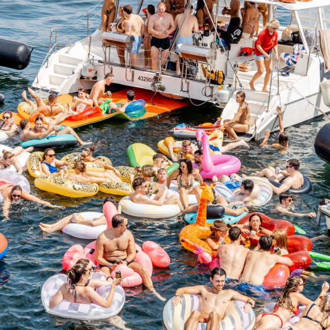A lively group of people enjoy a sunny day on colorful inflatable floats and swimming near a large catamaran boat anchored on the water, with more people socializing on the boat deck.