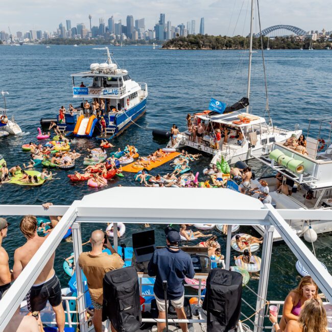 A lively scene on Sydney Harbour shows partygoers on boats and colorful inflatables, with a DJ playing music, city skyline and Sydney Harbour Bridge visible in the background. People are swimming and dancing in the sun.
