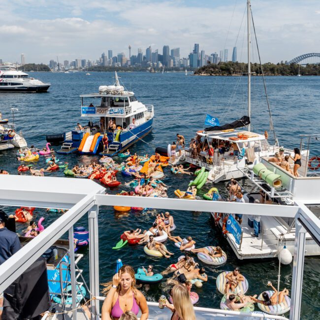 A lively scene of people on boats and inflatable floats enjoying a party on the water with the Sydney city skyline and Harbour Bridge visible in the background under a partly cloudy sky.