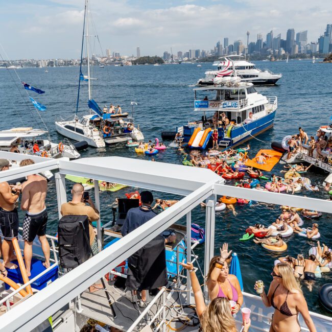 A lively boat party on a sunny day, with people in swimsuits on yachts and inflatables in the water, and the city skyline visible in the background.