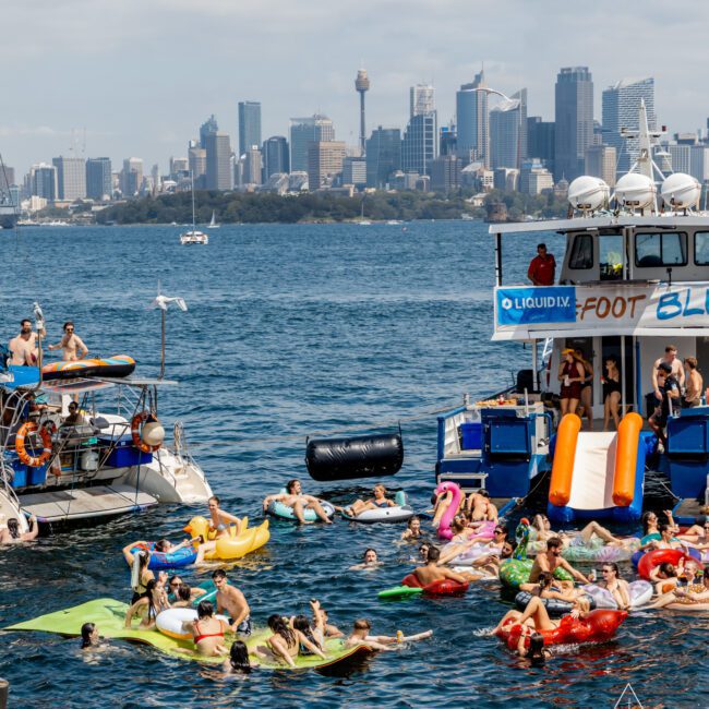 People relax on colorful inflatables in the water near boats during a sunny day, with the Sydney city skyline visible in the background. A slide leads from one boat into the harbor.