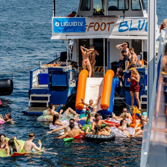 A lively group of people enjoy a party around a boat with a slide on the water. Some are swimming or floating on inflatables, while others relax or dance on the boat under sunny, clear weather.