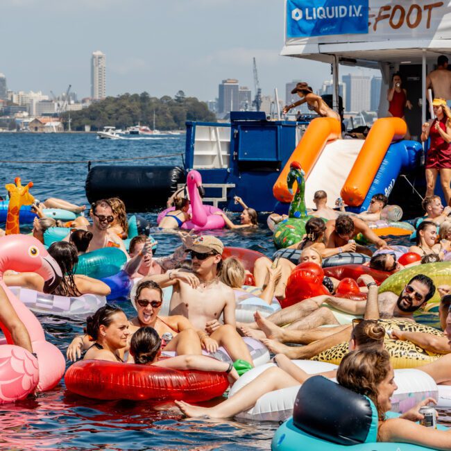 A lively group of people relax on colorful inflatable pool floats in the water beside a large boat on a sunny day, with city buildings and trees visible in the background.