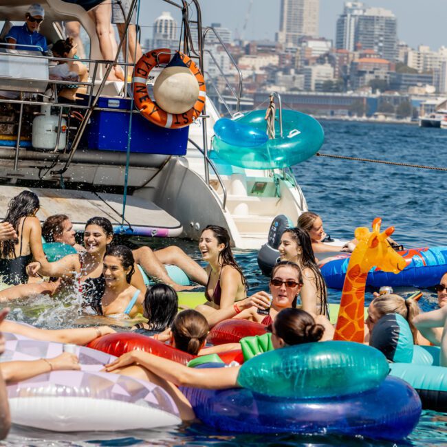 A group of people enjoy a pool party on the sea, floating on colorful inflatables near a yacht, with a city skyline in the background under a sunny sky.