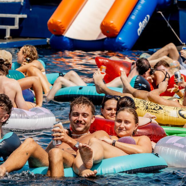 A group of people relax on colorful inflatable tubes in the water near a boat with an orange slide. Most are smiling, holding drinks, and enjoying the sunny day. The scene is lively and cheerful.