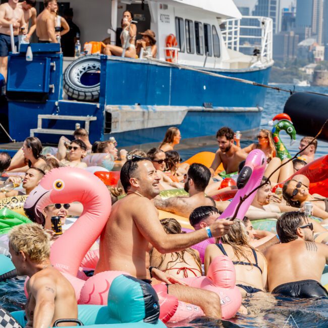A crowd of people in swimsuits float on colorful inflatable pool toys, including flamingos, near a large blue and white boat on the water in a city harbor on a sunny day.