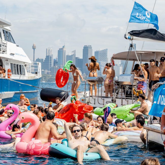 A lively boat party with many people in swimsuits enjoying the water on colorful inflatable floats, with boats and the city skyline in the background under a partly cloudy sky.