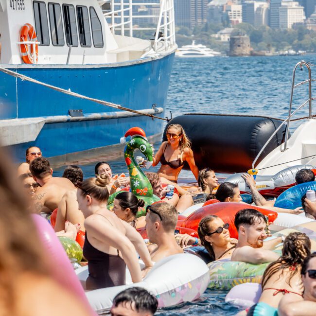 A crowd of people on colorful pool floats and inflatables gather in the water next to a docked boat, with city buildings visible in the background on a sunny day.