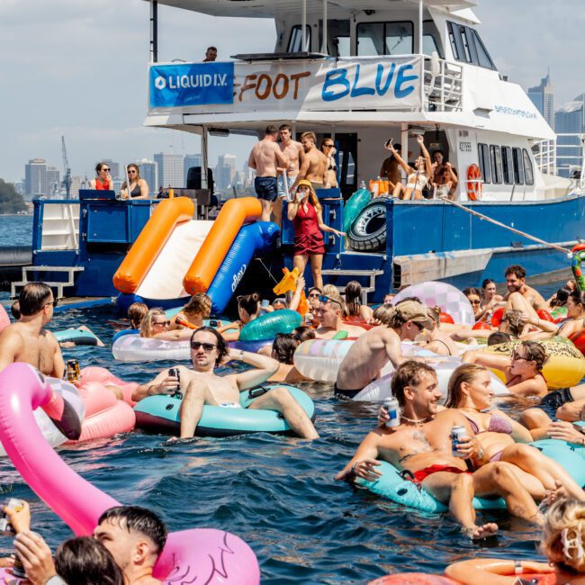 A lively pool party with people relaxing on colorful inflatables in the water near a blue and white boat. Some are using the boat’s orange slides while others enjoy drinks under sunny skies. City buildings are visible in the background.
