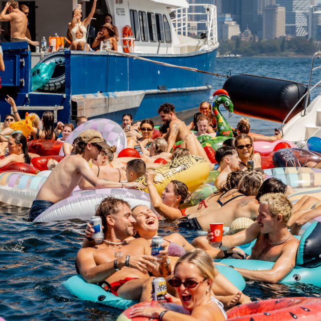 A lively group of people relax in colorful inflatable tubes on the water, enjoying drinks and sunlight near docked boats, with a city skyline in the background.