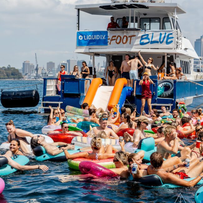 A large group of people relax on colorful inflatables in the water near a boat with a slide. The boat has a "FOOT BLUE" sign, and the city skyline is visible in the background. The atmosphere is lively and festive.