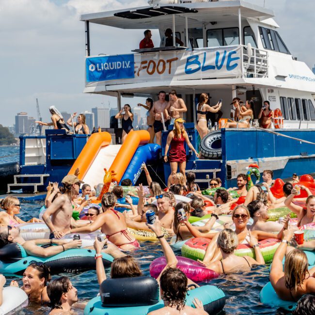 A large group of people in colorful pool floats enjoy a lively party in the water near a yacht, which has slides and a "Foot Blue" banner. Sunny weather and city buildings are visible in the background.