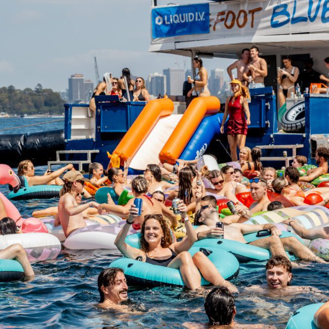 A crowd of people relax on colorful pool floats, holding drinks and smiling in the water near a boat with waterslides. The boat has a “LIQUID” banner and people are standing, watching, and swimming around.