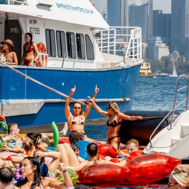 A group of people in swimsuits relax on colorful inflatable floats in the water near a blue and white boat, with a city skyline and tall buildings visible in the background. Two women stand and pose on a boat ladder.