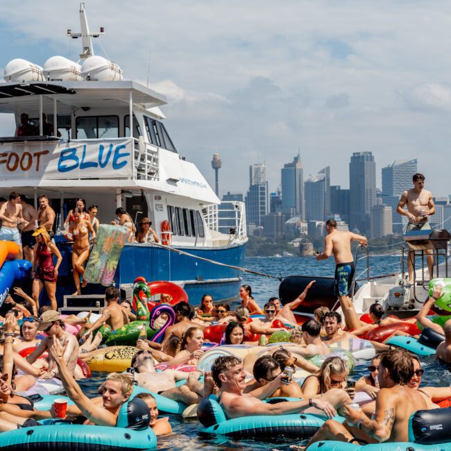 A large group of people on inflatable floats enjoy a lively party in the water near a boat named "Footloose Blue," with a city skyline in the background under a clear sky.