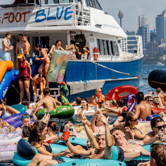 A large group of people in colorful pool floats and inflatables celebrate in the water near a blue and white yacht, with a city skyline in the background under a clear sky.