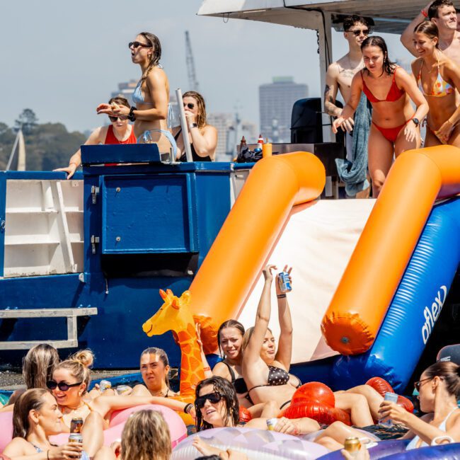 A crowd of people in swimsuits enjoy a pool party, lounging on colorful inflatables near a boat with a "LIQUID I.V." banner. Some are taking photos and smiling, with a city skyline in the background.