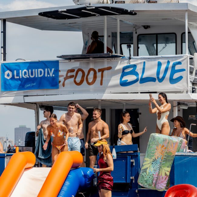 A group of people in swimsuits enjoy a pool party on a boat named "Foot Blue," with pool floats and inflatable toys, under a sunny sky. A "Liquid I.V." banner is displayed on the upper deck.