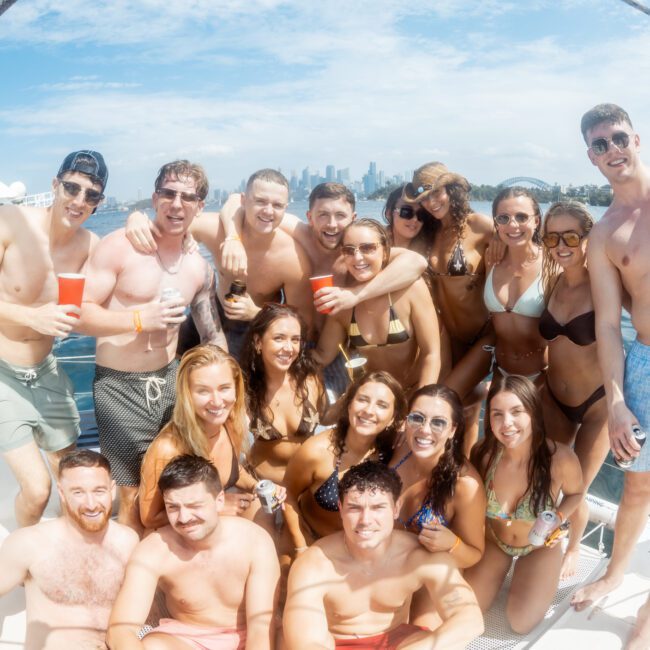 A group of young adults in swimsuits smile and pose together on a boat in sunny weather, with water, other boats, and a city skyline visible in the background.
