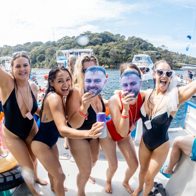 A group of women in swimsuits smile and pose on a boat, holding drinks and large cutout face masks. Other boats and people are visible on the water, with a wooded shoreline in the background.