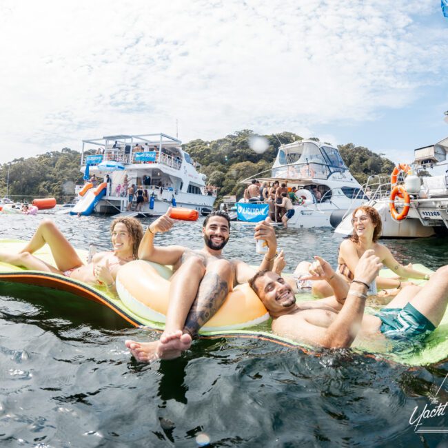 Four young adults relax and smile on a float in the water, holding drinks, surrounded by boats and other people at a lively yacht party under a partly cloudy sky.