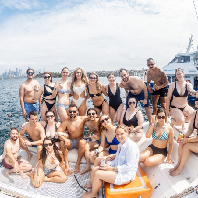 A large group of people in swimsuits pose and smile on a sailboat under sunny skies, with water and a city skyline in the background. Some hold drinks, and everyone appears to be enjoying a summer day.