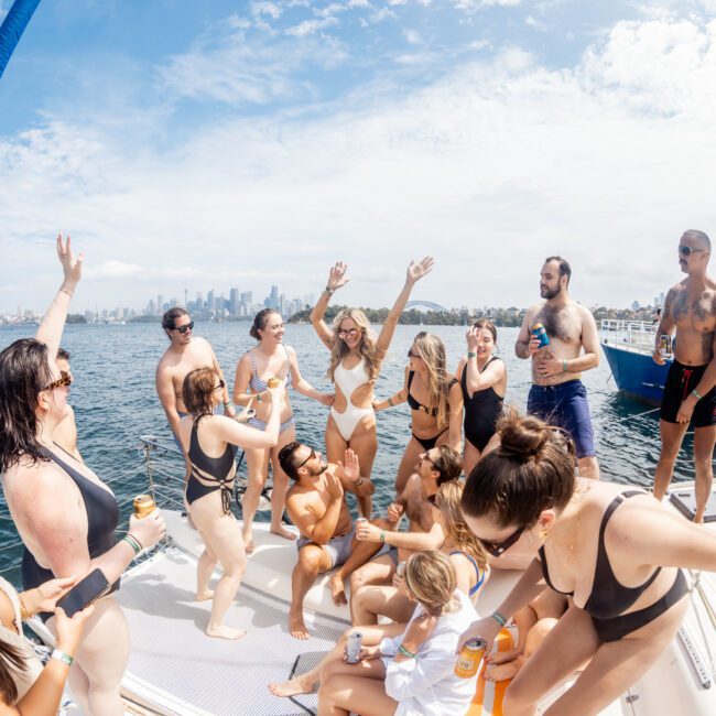 A group of people in swimsuits laugh, dance, and pose together on a boat under a partly cloudy sky, with a city skyline and another boat visible in the background. The scene is joyful and energetic.