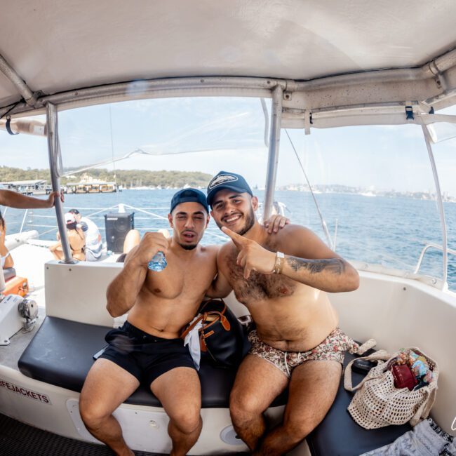 Two men in swimwear sit together on a boat, smiling and posing with arms around each other. Clear blue water and distant shoreline are visible in the background. Another person drinks water on the left. A logo reads "Yacht Social Club.