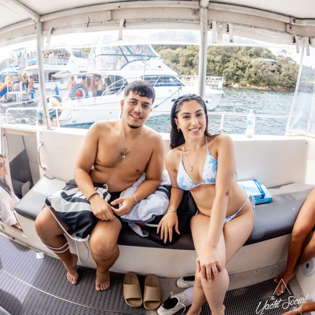 A man and woman in swimwear sit smiling on a boat bench, with other yachts and people visible on the water in the background. It is a sunny day, and the scene looks relaxed and cheerful.