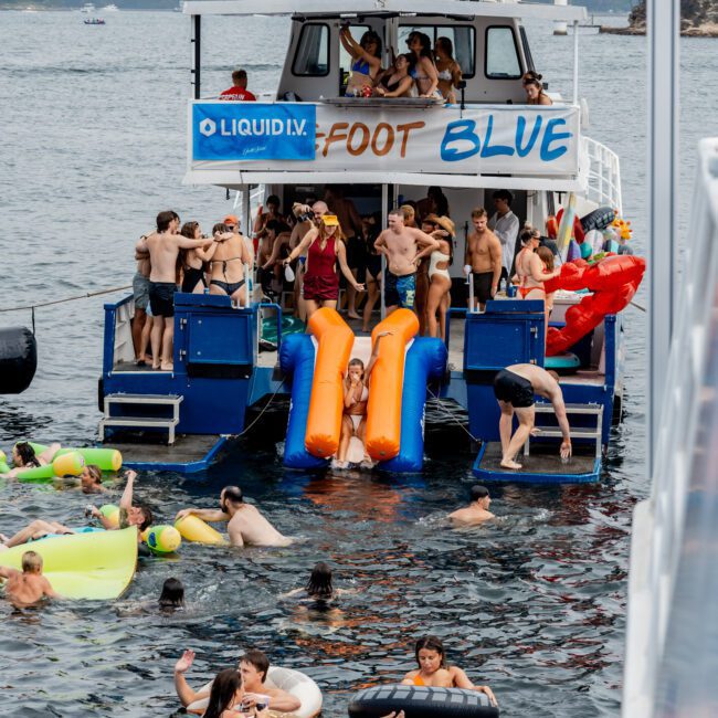 A crowded party boat with slides is anchored on a city waterfront. People in swimsuits are dancing on the deck, entering the water, and relaxing on colorful pool floats, while tall buildings are visible in the background.