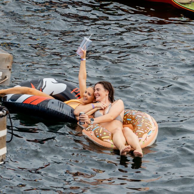 Two women relax on inflatable pool floats, one shaped like a donut and the other a black creature, smiling and holding drinks in the water; a man lounges on a floating mat in the background.