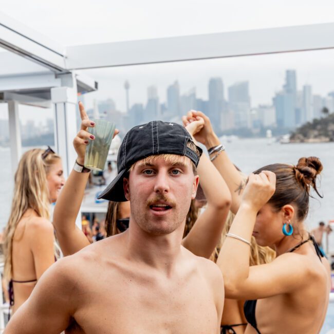 A shirtless man wearing a backwards cap stands in front of dancing people at an outdoor pool party, with a city skyline and water in the background on a cloudy day.