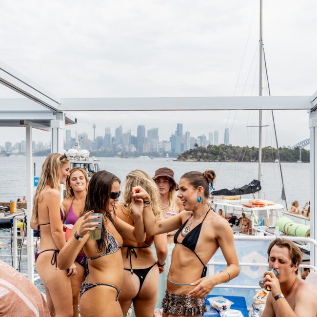 A group of young women in swimsuits dance and enjoy drinks on a boat with more people relaxing in the background. The city skyline and Sydney Harbour Bridge are visible across the water under a cloudy sky.