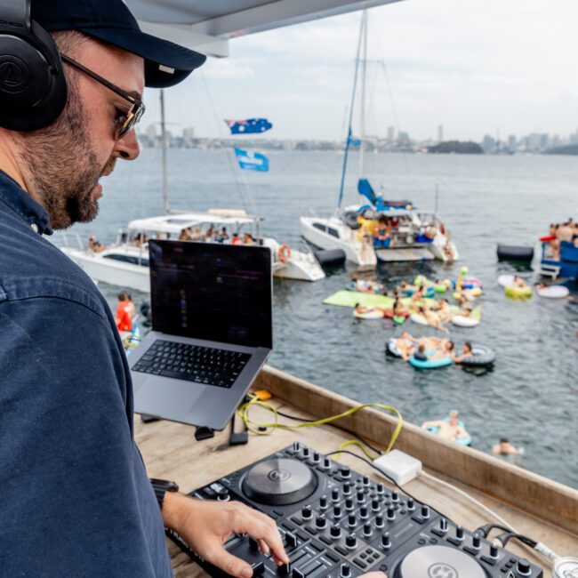 A DJ wearing headphones mixes music on a deck overlooking a lively harbor, where people on boats and inflatables gather for a party on the water.