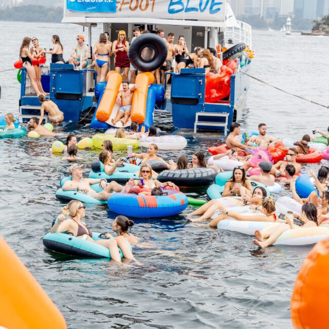 A large group of people relax on colorful floaties around a boat labeled "Foot Blue" during a lively party on the water, with a city skyline visible in the background.