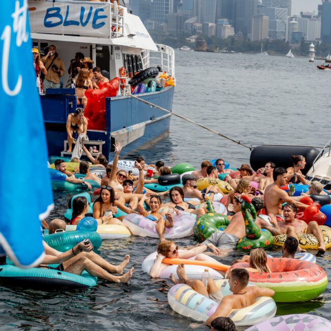 Dozens of people in colorful inflatable pool floats party in the water next to a boat labeled "BLUE," with a city skyline in the background. The scene is lively with music, laughter, and summer fun.